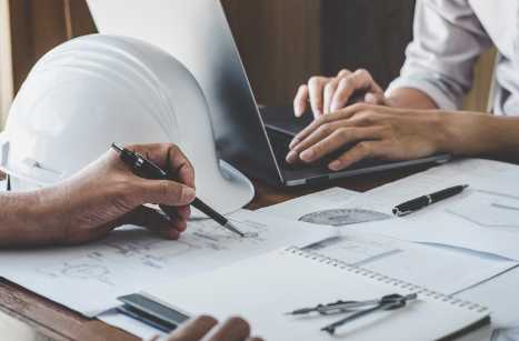 A laptop and hardhat sit on a table where people are planning a roof installation.